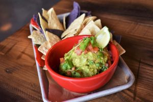 Dos Gatos Appetizer Menu guacamole in a red bowl on a tray with tri-colored tortilla chips 